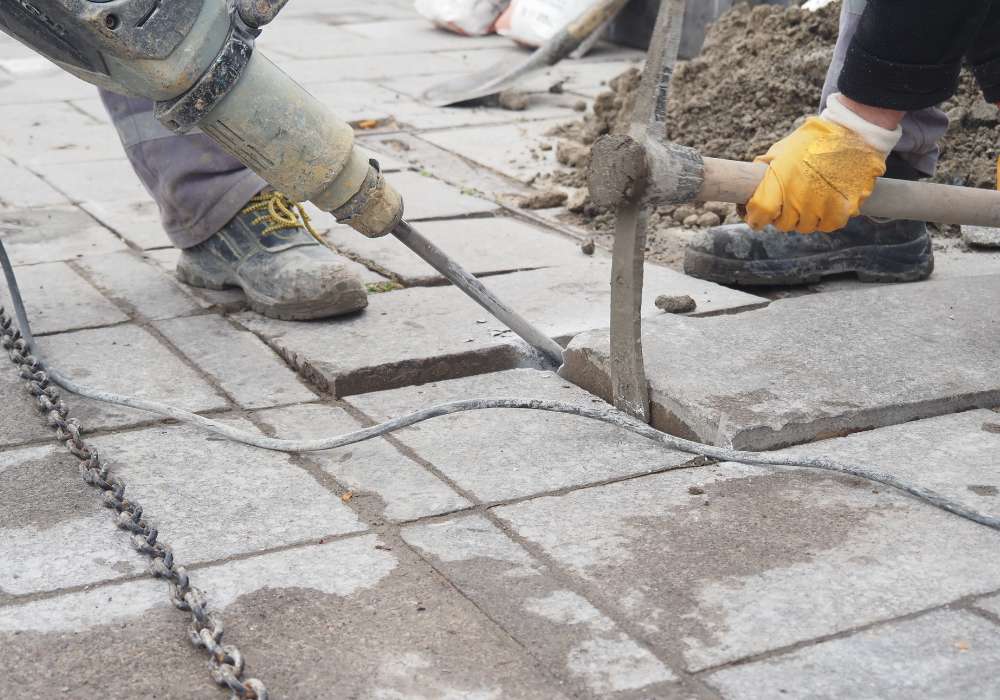 Worker leveling an uneven concrete sidewalk slab outside a New York City brownstone using slab-lifting equipment.